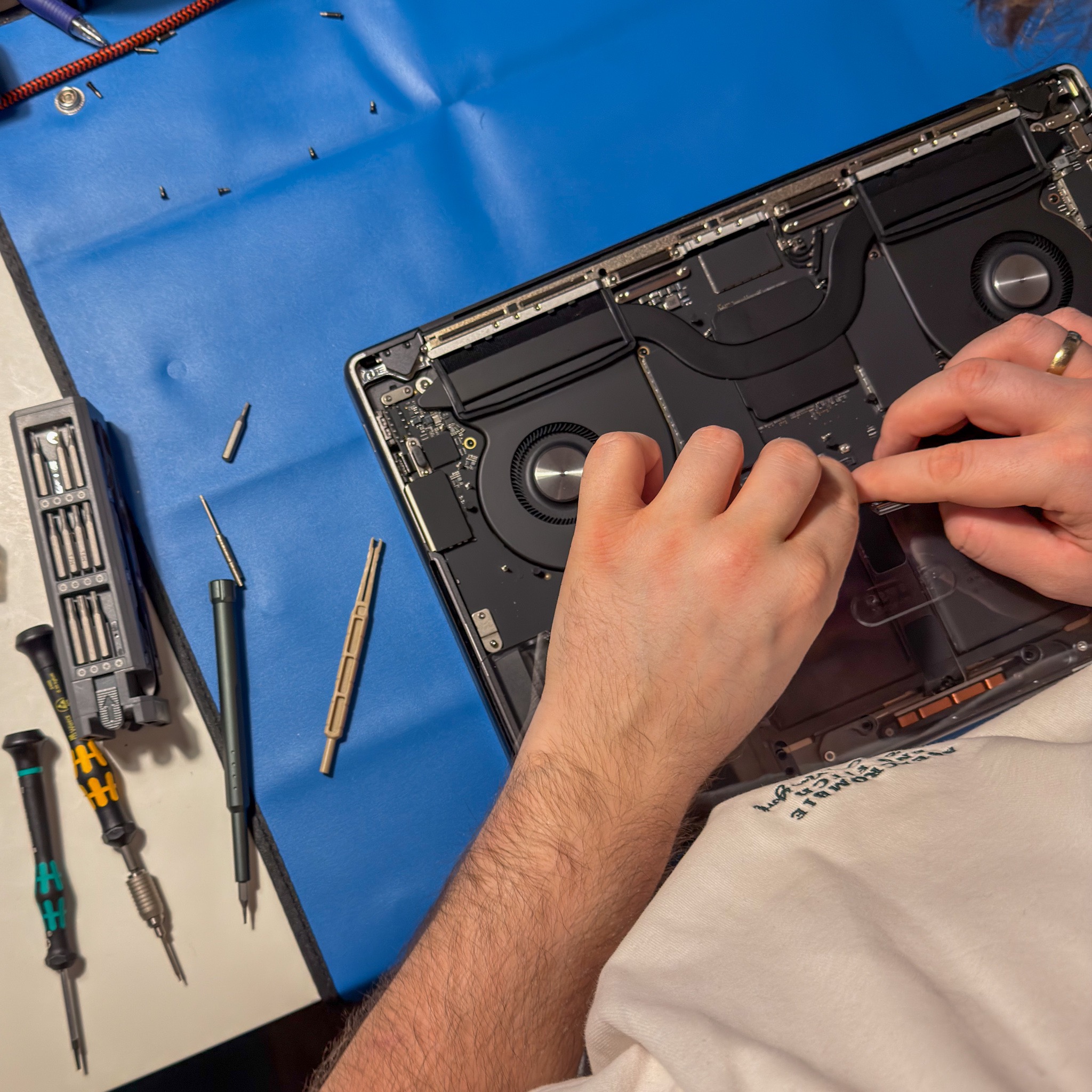 Otwarty laptop na niebieskiej macie serwisowej, narzędzia obok. Dłonie serwisanta w trakcie naprawy, widoczne elementy wewnętrzne komputera.