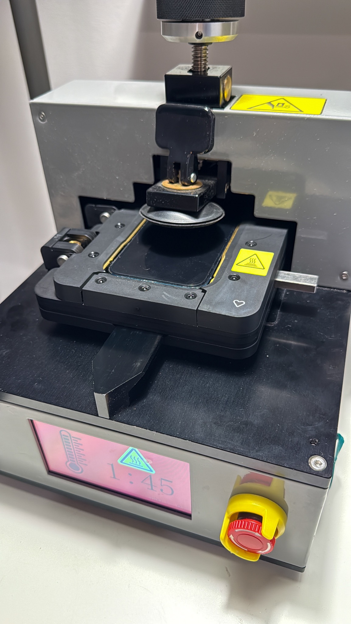 Specjalistyczna maszyna do naprawy telefonów w trakcie pracy. Widoczny ekran z temperaturą i czasem. Żółte naklejki ostrzegawcze. Precyzyjne narzędzie serwisowe.