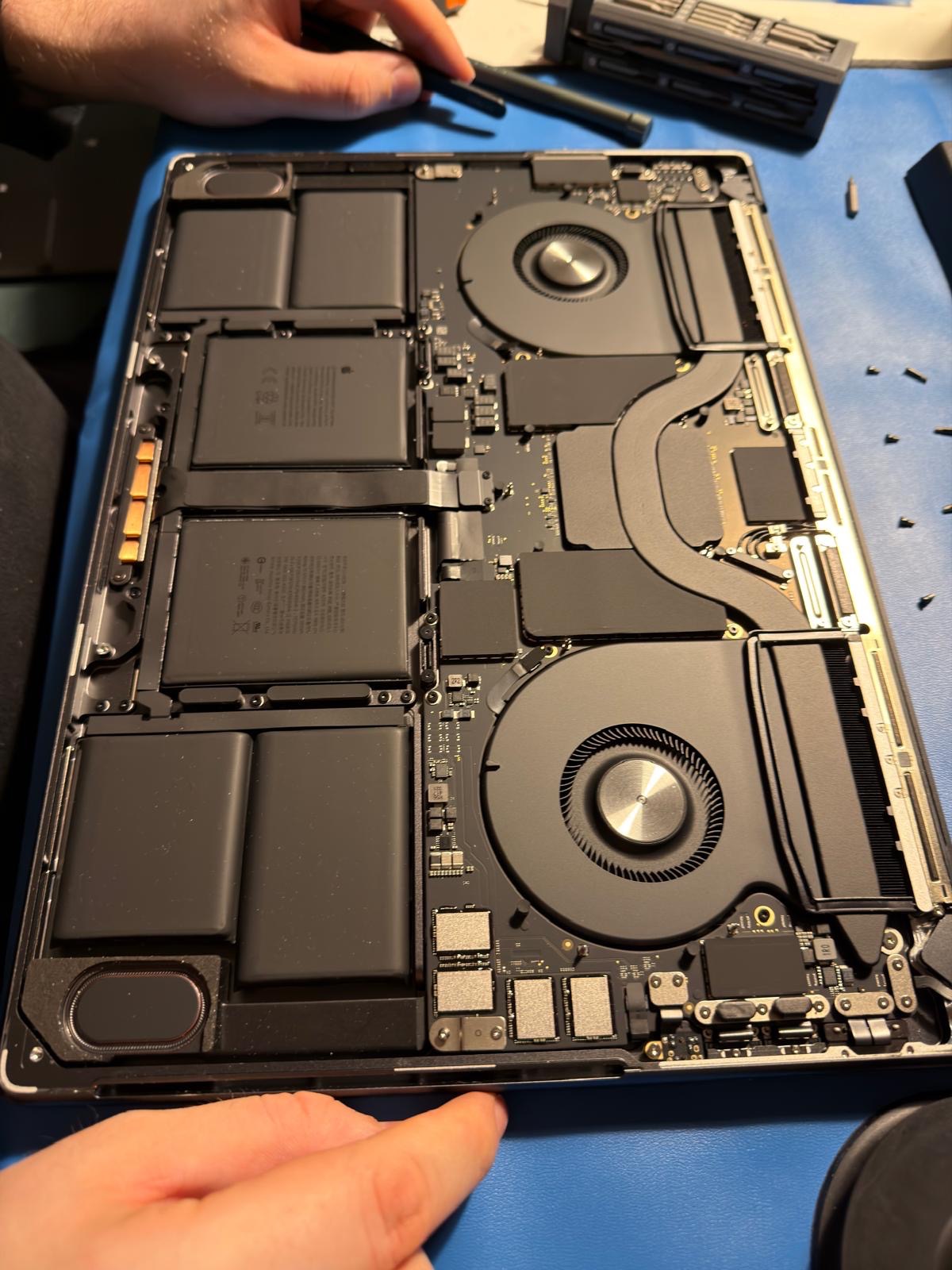 Otwarty laptop MacBook Pro z widocznymi bateriami, wentylatorami i płytą główną. Widoczne narzędzia i dłonie osoby serwisującej. Detaliczne ujęcie wnętrza urządzenia.