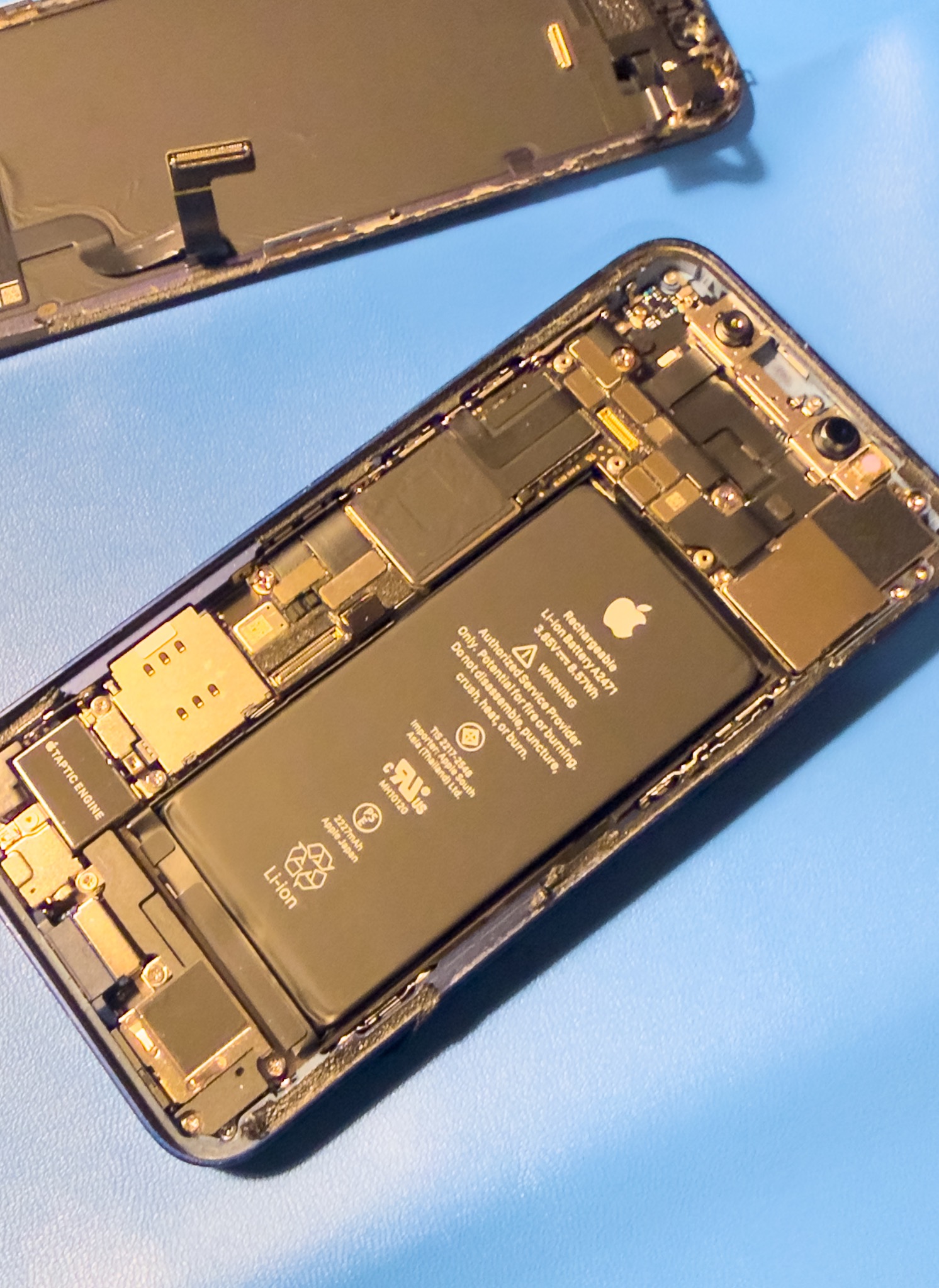 Otwarty iPhone z widoczną baterią i wewnętrznymi komponentami na niebieskim tle. Widoczne ślady demontażu, sugerujące serwis lub naprawę telefonu.