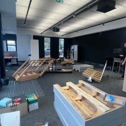 SpeedTrade - Demontaż i przygotowanie do transportu ekspozytorów schodów w biurze. Widoczne elementy konstrukcyjne, palety i kartony. Oświetlenie sufitowe, czarne ściany w tle.