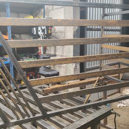 Marcintech - Konstrukcja metalowa w warsztacie: spawana rama z profili, widoczne narzędzia i regały. Surowy, industrialny klimat. Elementy balustrady lub ogrodzenia w trakcie tworzenia.