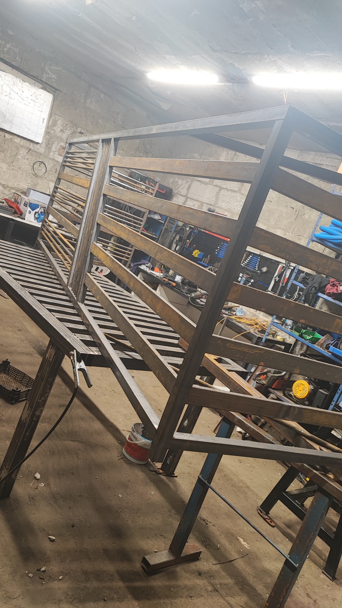 Konstrukcja metalowa w warsztacie, spawana z profili stalowych, widoczne narzędzia i elementy wyposażenia warsztatu w tle. Surowy stan przed wykończeniem.