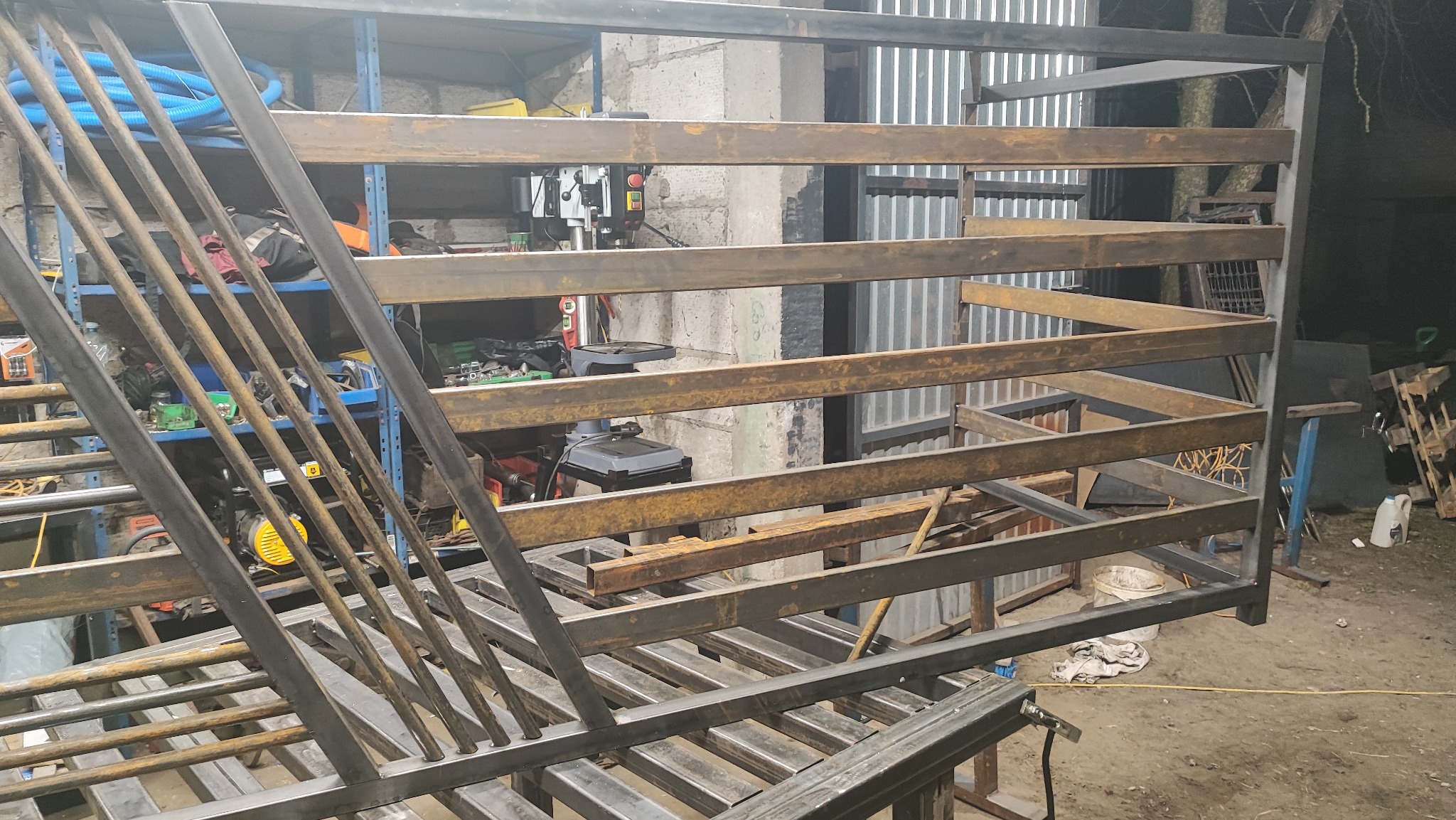 Konstrukcja metalowa w warsztacie: spawana rama z profili, widoczne narzędzia i regały. Surowy, industrialny klimat. Elementy balustrady lub ogrodzenia w trakcie tworzenia.