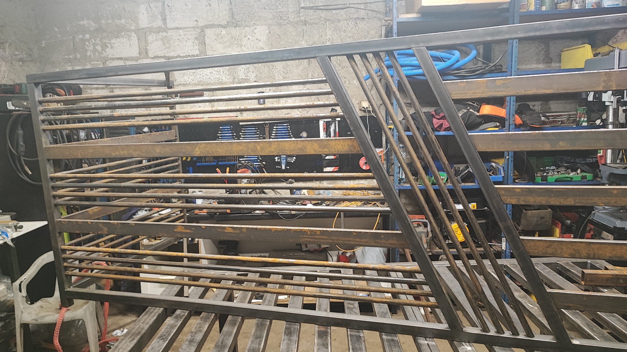 Surowa, metalowa konstrukcja kratowa w warsztacie, z widocznymi spawami i elementami w trakcie montażu. Tło stanowią narzędzia i materiały warsztatowe.