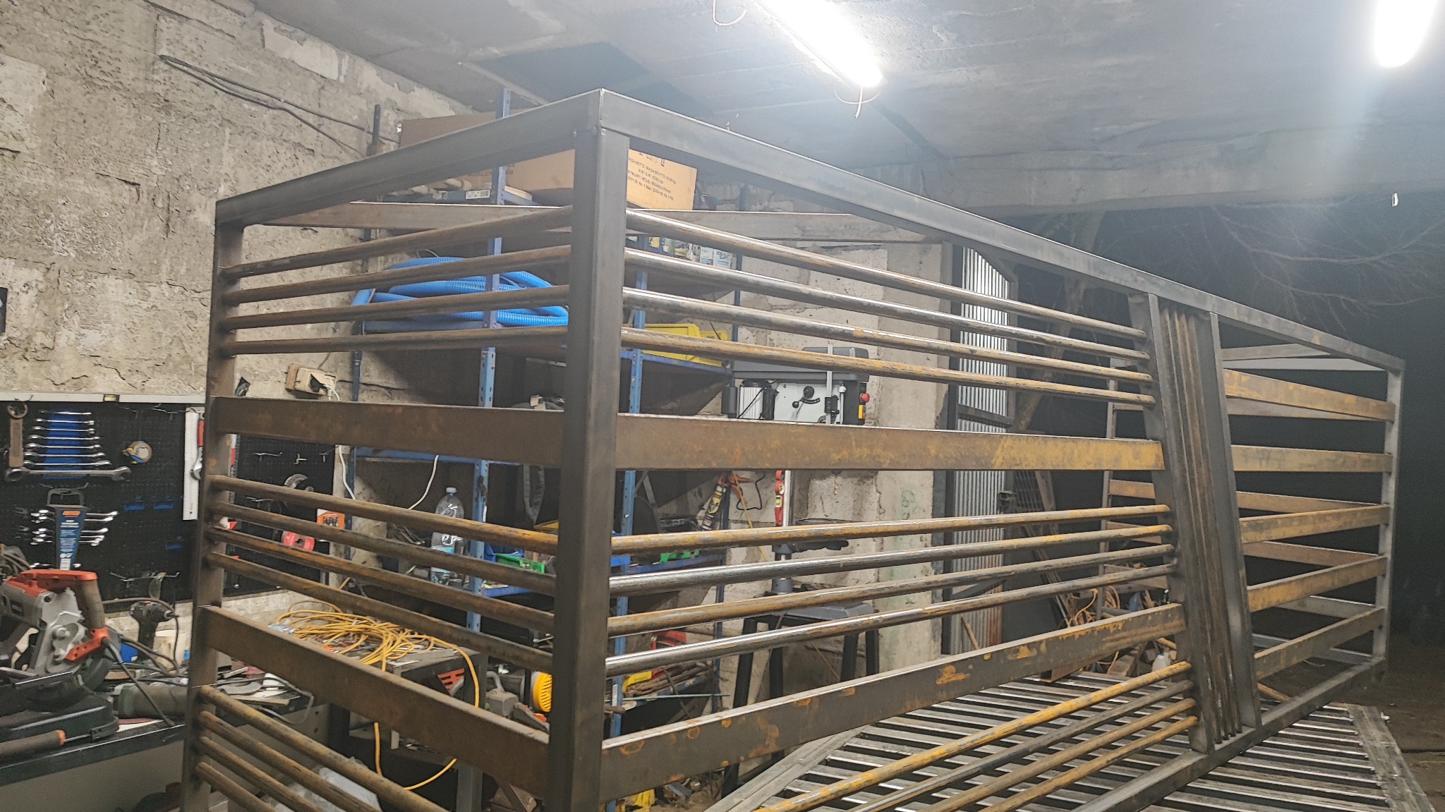 Surowa, metalowa konstrukcja z poziomych prętów w warsztacie, prawdopodobnie fragment bramy lub ogrodzenia, z widocznymi narzędziami i elementami wyposażenia.