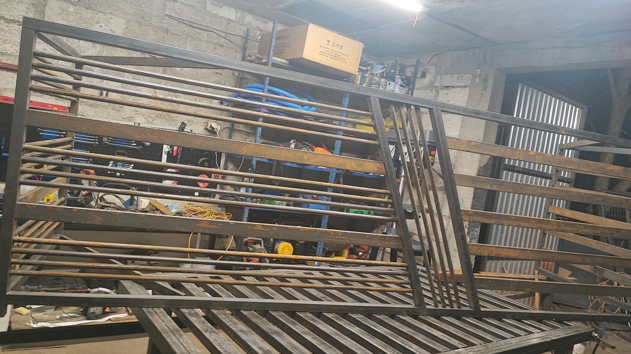 Surowa, metalowa konstrukcja bramy wjazdowej w warsztacie. Widoczne spawy i elementy konstrukcyjne. Tło stanowią narzędzia i materiały warsztatowe.