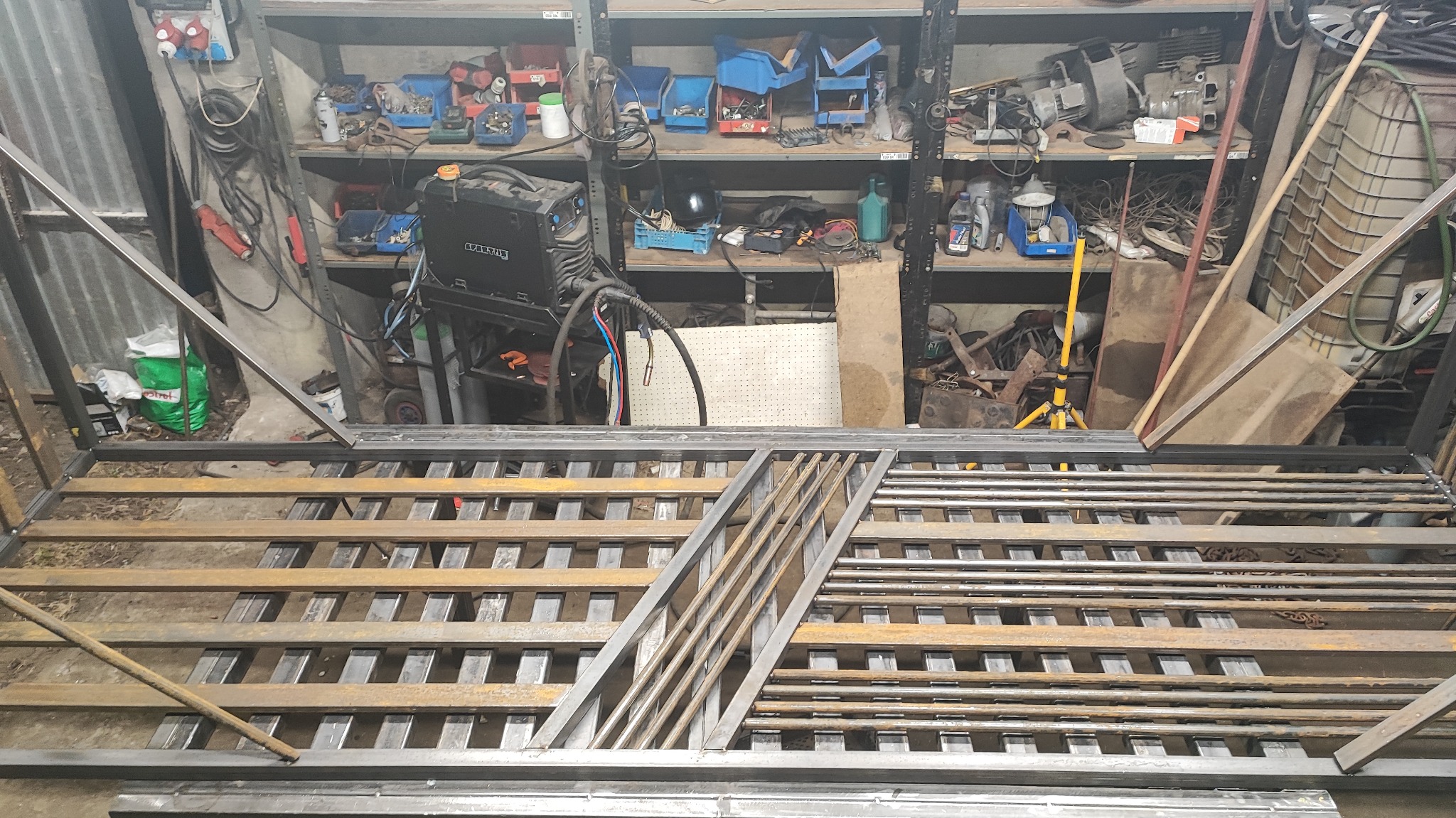 Metalowa konstrukcja w warsztacie, prawdopodobnie fragment bramy lub ogrodzenia, w trakcie spawania. Widoczne narzędzia i materiały.