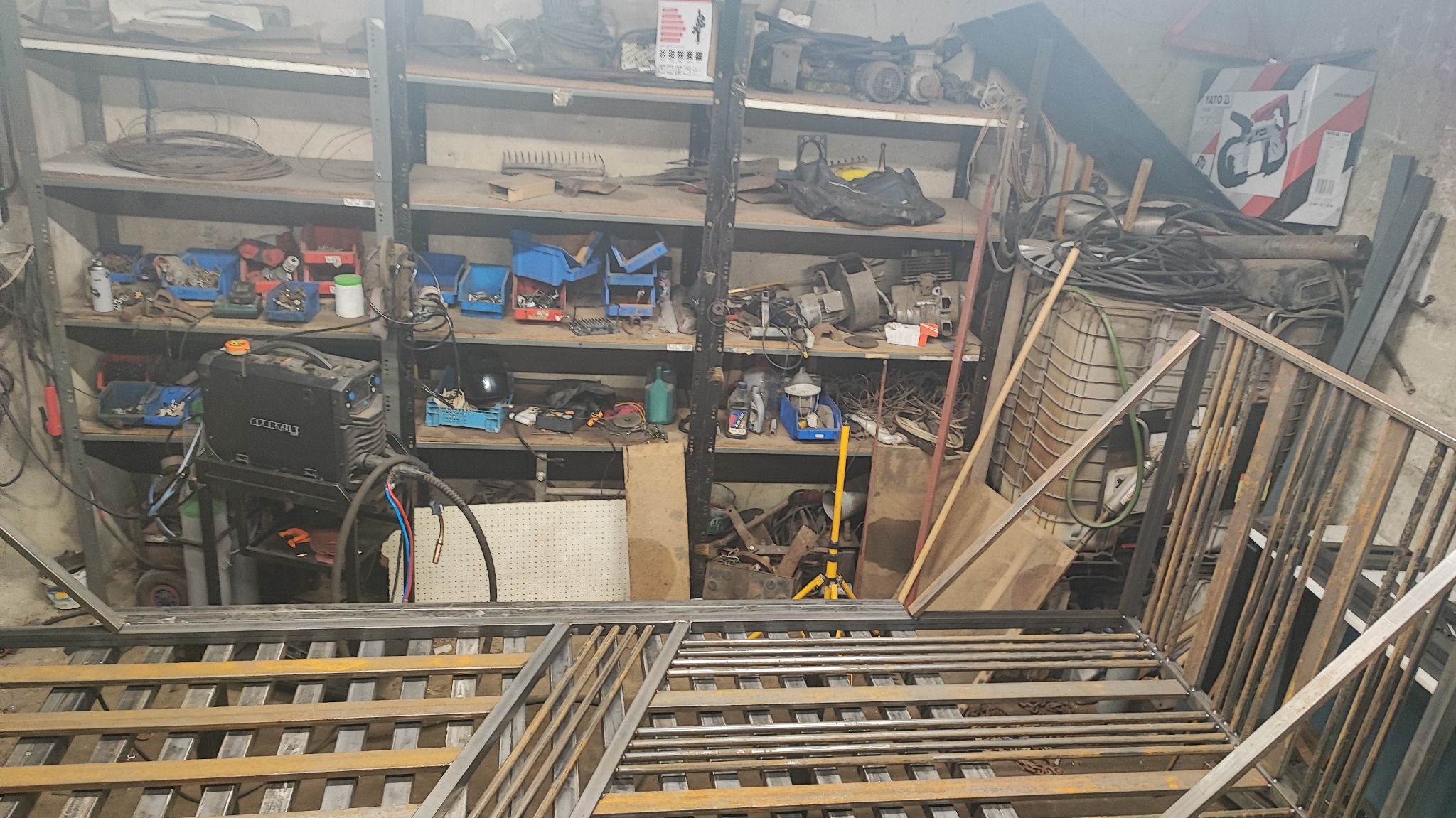 Metalowa konstrukcja w warsztacie, widoczne półki z narzędziami i spawarka. Elementy konstrukcyjne w trakcie tworzenia, surowy wygląd.