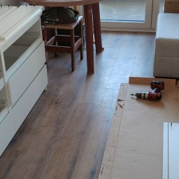 Jacek Sankowski - Biała komoda z szufladami w trakcie montażu, widoczna wiertarka i elementy meblowe na kartonie, drewniana podłoga i fragment sofy w tle. Ujęcie z perspektywy.