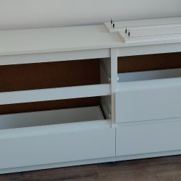 Jacek Sankowski - Biała komoda z szufladami w trakcie montażu. Częściowo zmontowana, widoczne prowadnice szuflad i elementy do skręcania na blacie. Ujęcie z boku, na drewnianej podłodze.