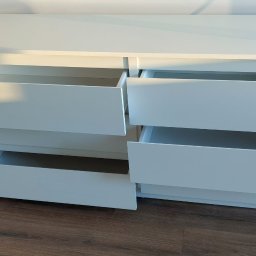 Jacek Sankowski - Biała komoda z sześcioma szufladami, z których trzy są otwarte, ukazując puste wnętrze. Komoda stoi na drewnianej podłodze przy białej ścianie.