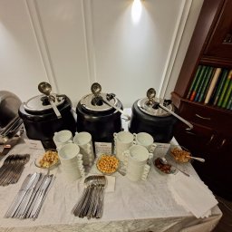 Maincourse.robertpawlina - Bufet z trzema zupami krem w podgrzewaczach, stosami białych filiżanek, orzechami i grzankami na stole z białym obrusem. W tle regał z książkami.