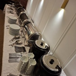 Maincourse.robertpawlina - Stół cateringowy z podgrzewaczami, stosem talerzy, sztućcami i filiżankami. Biały obrus z delikatnym wzorem, oświetlenie punktowe na ścianie w tle.