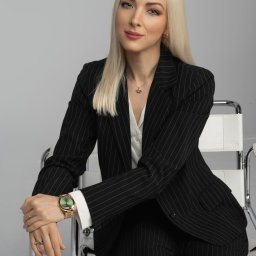 Kancelaria Adwokacka Adwokat Karolina M. Stempniewska - Portret kobiety w czarnym garniturze w prążki, siedzącej na białym krześle. Złoty zegarek i naszyjnik z koroną dodają elegancji. Profesjonalny wizerunek.