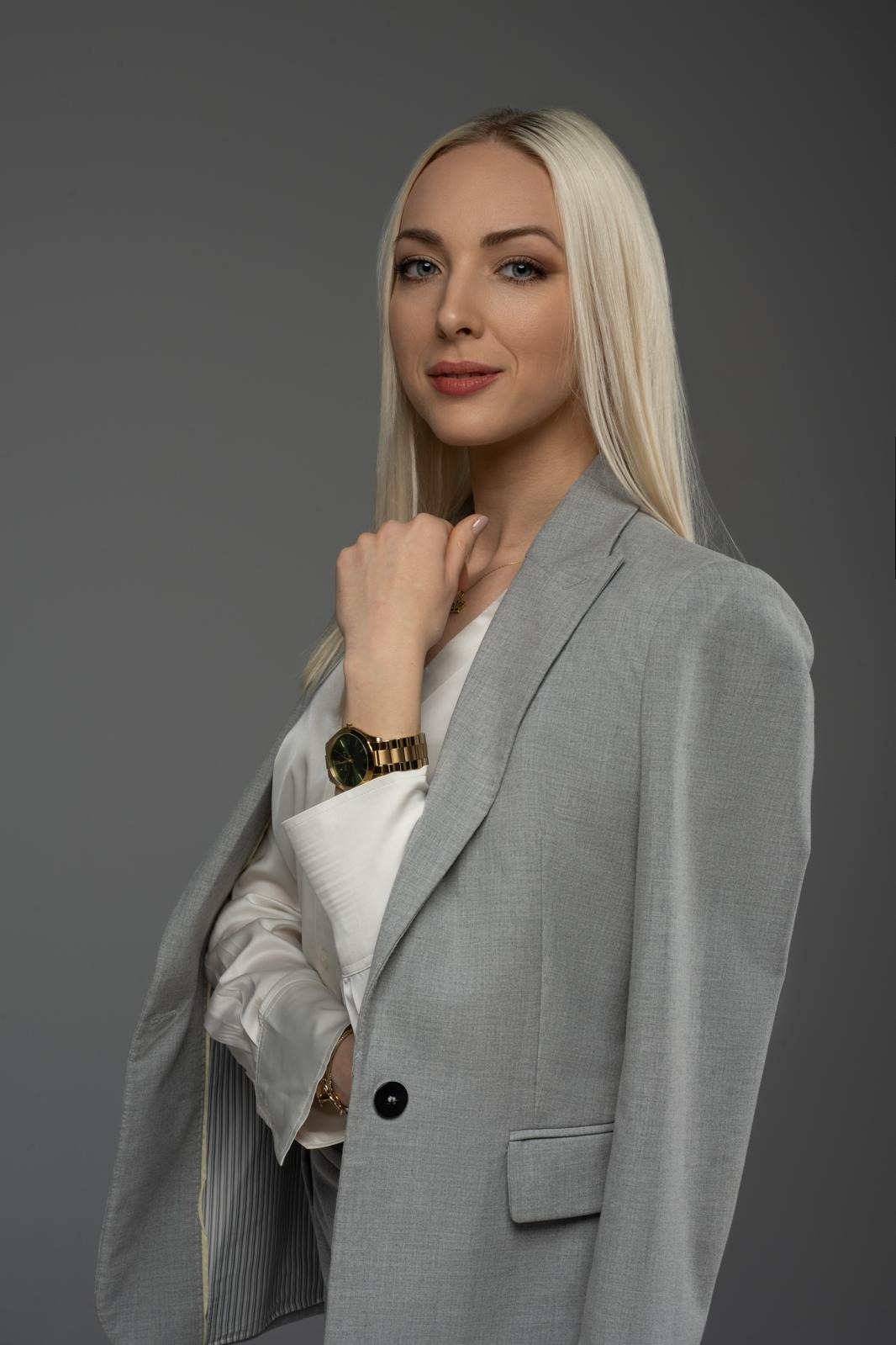 Portret kobiety w szarym żakiecie i białej koszuli, z blond włosami i złotym zegarkiem, na jednolitym szarym tle. Profesjonalny wizerunek.