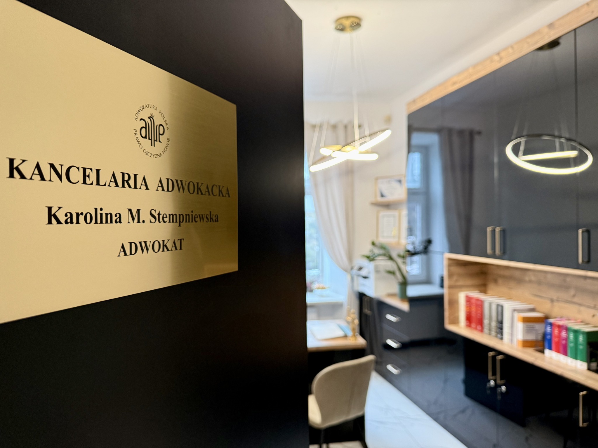 Złota tabliczka z napisem 'Kancelaria Adwokacka Karolina M. Stempniewska Adwokat' na czarnej ścianie, widok na wnętrze biura z nowoczesnym oświetleniem i regałem z książkami.