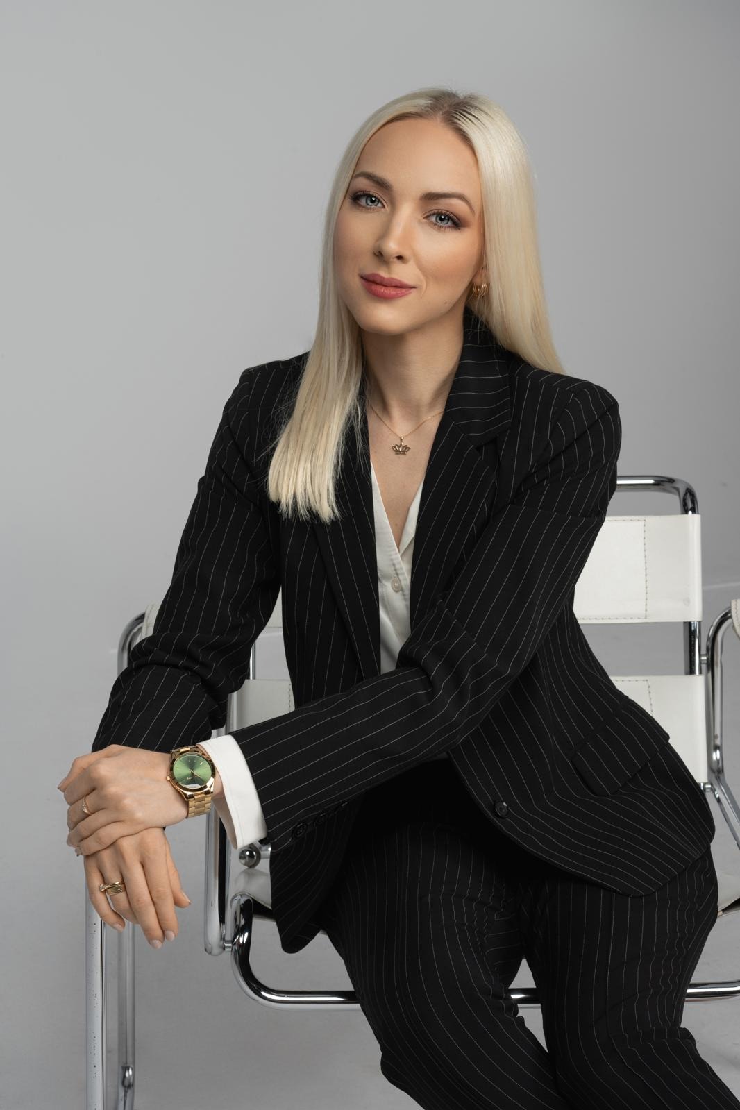 Portret kobiety w czarnym garniturze w prążki, siedzącej na białym krześle. Złoty zegarek i naszyjnik z koroną dodają elegancji. Profesjonalny wizerunek.