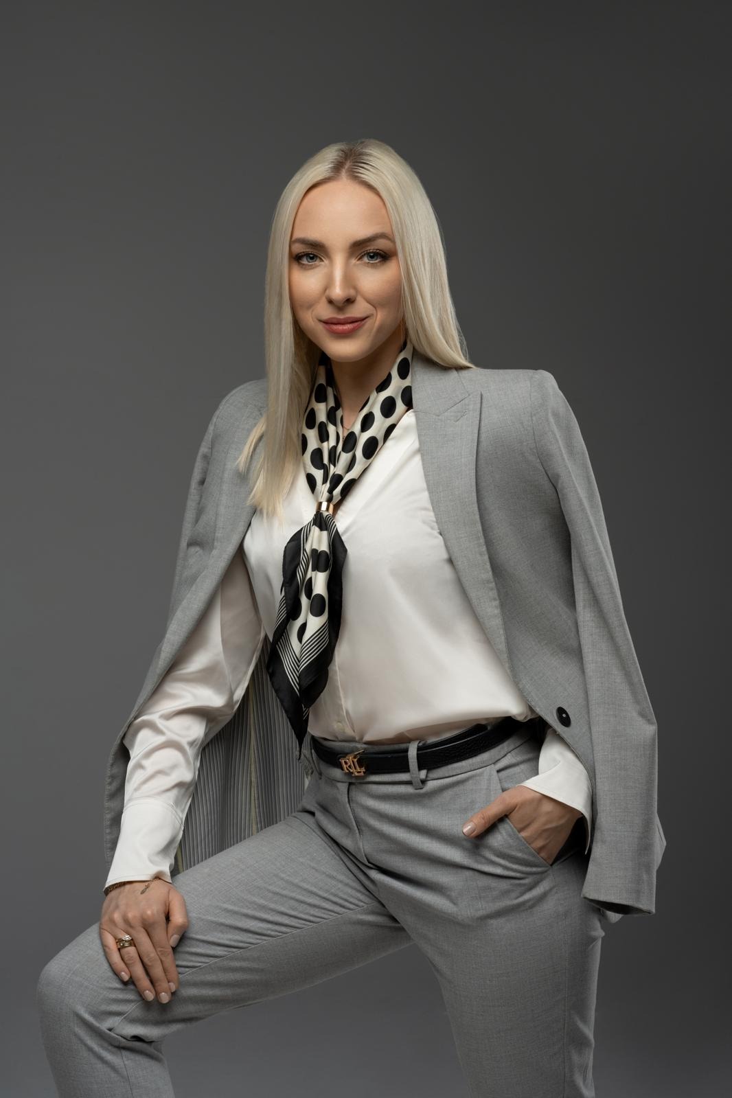 Portret kobiety w szarym garniturze i bluzce z apaszką w kropki, pozującej na szarym tle. Uśmiech, profesjonalny wygląd, dłoń w kieszeni spodni.