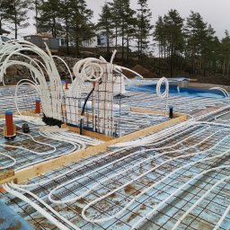 NORMEST - Instalacja ogrzewania podłogowego w domu szkieletowym: białe rury ułożone na izolacji, widoczne elementy konstrukcji i zbrojenia. Tło stanowią drzewa i budynek w budowie.