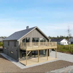 NORMEST - Nowoczesny dom szkieletowy z dużym drewnianym balkonem i szarą elewacją. Widok z zewnątrz, w tle inne domy i zieleń. Teren wokół domu przygotowany pod zagospodarowanie.