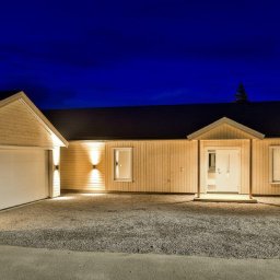 NORMEST - Nowoczesny dom szkieletowy z garażem oświetlony zewnętrznymi lampami ściennymi, widok nocą na tle ciemnego nieba.