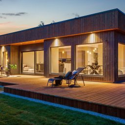 NORMEST - Nowoczesny dom z drewnianą elewacją i dużymi oknami, oświetlony ciepłym światłem o zmierzchu. Taras z leżakiem i meblami ogrodowymi. Minimalistyczny design.