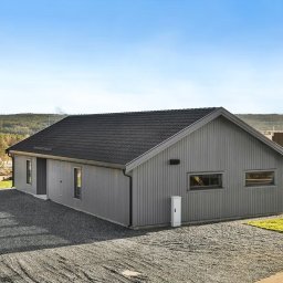 NORMEST - Nowoczesny, szary dom modułowy z ciemnym dachem na żwirowym podłożu, z widocznym fragmentem lasu w tle i elementami placu budowy obok.