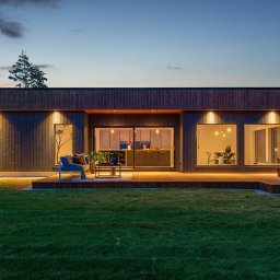 NORMEST - Nowoczesny dom modułowy z drewnianą elewacją i tarasem w Halden, oświetlony wieczornym światłem. Widoczne wnętrze przez duże okna, meble ogrodowe na tarasie.