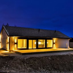 NORMEST - Oświetlony, parterowy dom z dużymi oknami i drewnianym tarasem, widziany z zewnątrz o zmierzchu. Nowoczesna architektura z elementami drewna i minimalistycznym designem.