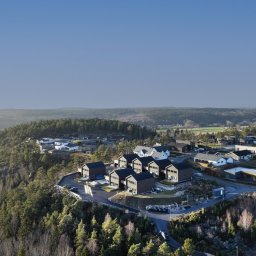 NORMEST - Panoramiczny widok z lotu ptaka na osiedle domów modułowych z ciemnego drewna, położone na wzgórzu porośniętym lasem iglastym, w tle rozległe tereny zielone i błękitne niebo.