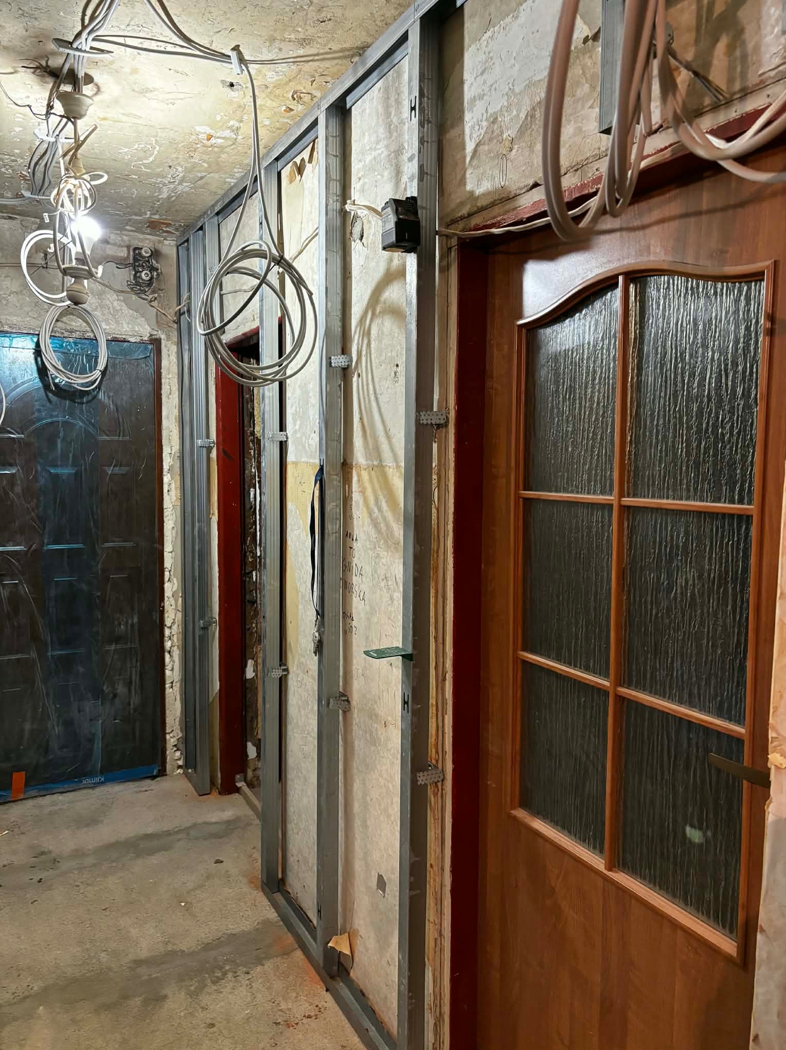 Remont wnętrza: metalowe profile ścian działowych, kable elektryczne zwisające z sufitu, widoczne drzwi z szybą ornamentową. Etap prac instalacyjnych.