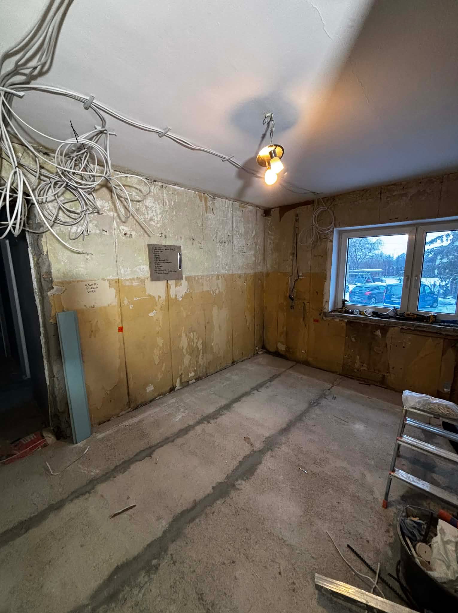 Remontowane wnętrze z odsłoniętymi przewodami elektrycznymi na suficie, surowymi ścianami i betonową podłogą. Widoczne narzędzia i drabina. Oświetlenie tymczasowe.