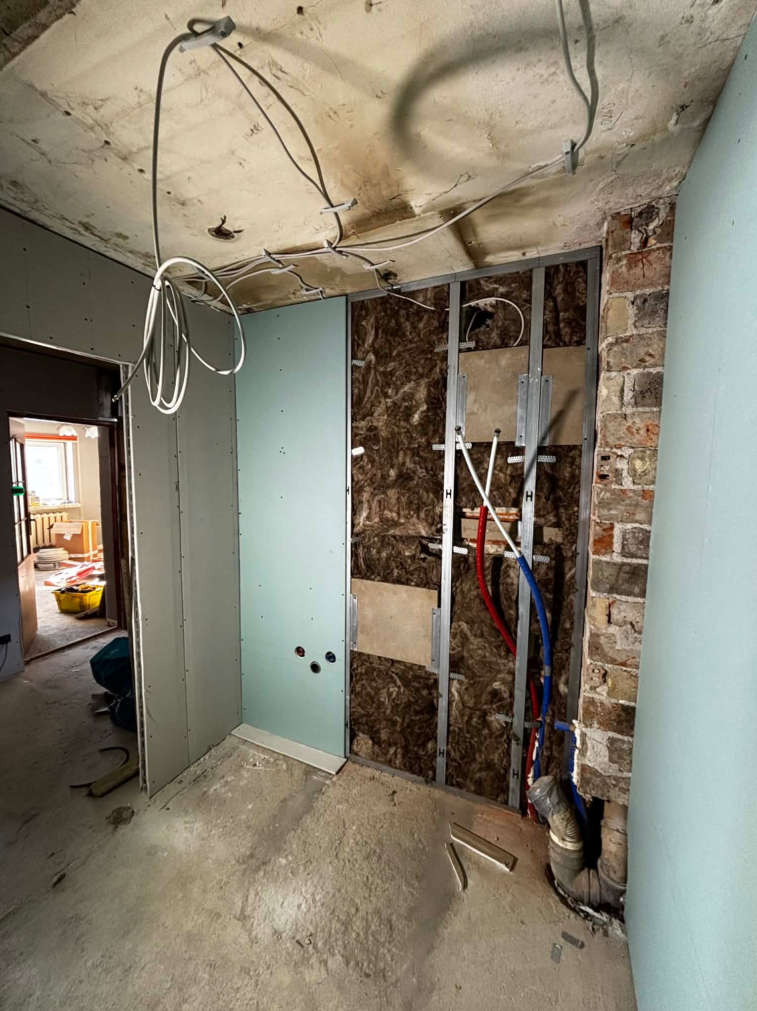 Remont łazienki w trakcie: widoczne instalacje (rury, kable), płyty gipsowe, izolacja. Surowy stan, przygotowanie do wykończenia. Fragment ściany z cegły.
