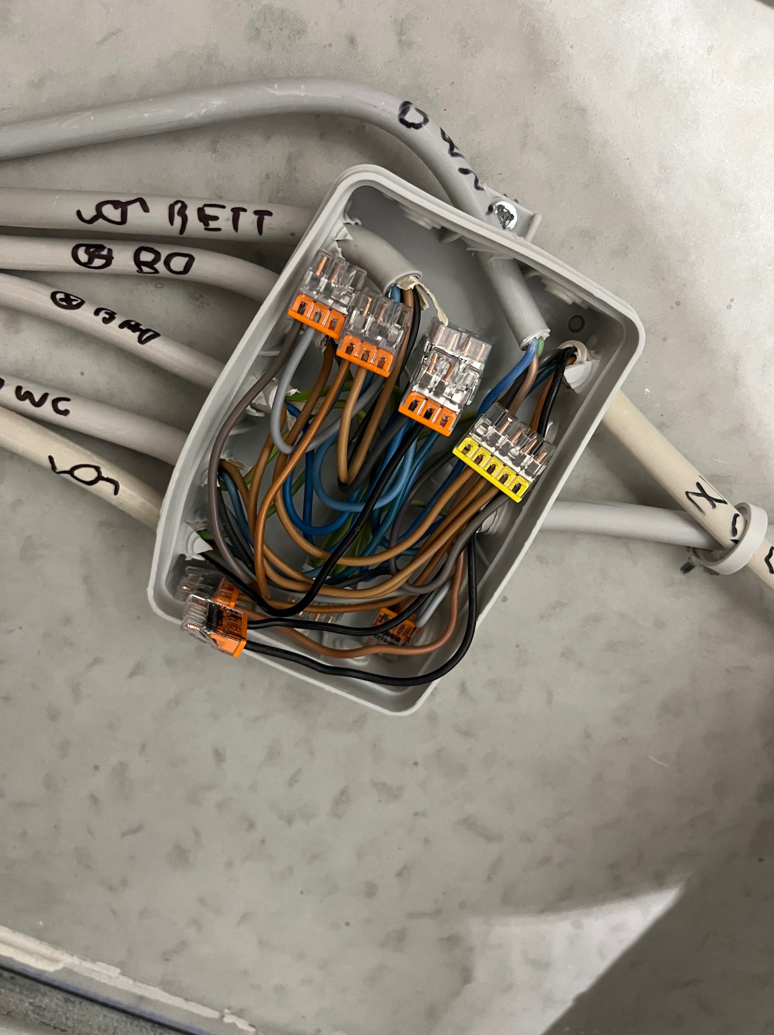 Szara puszka elektryczna z podłączonymi przewodami za pomocą złączek WAGO. Przewody opisane markerem, ułożone wzdłuż ściany. Widok z góry.