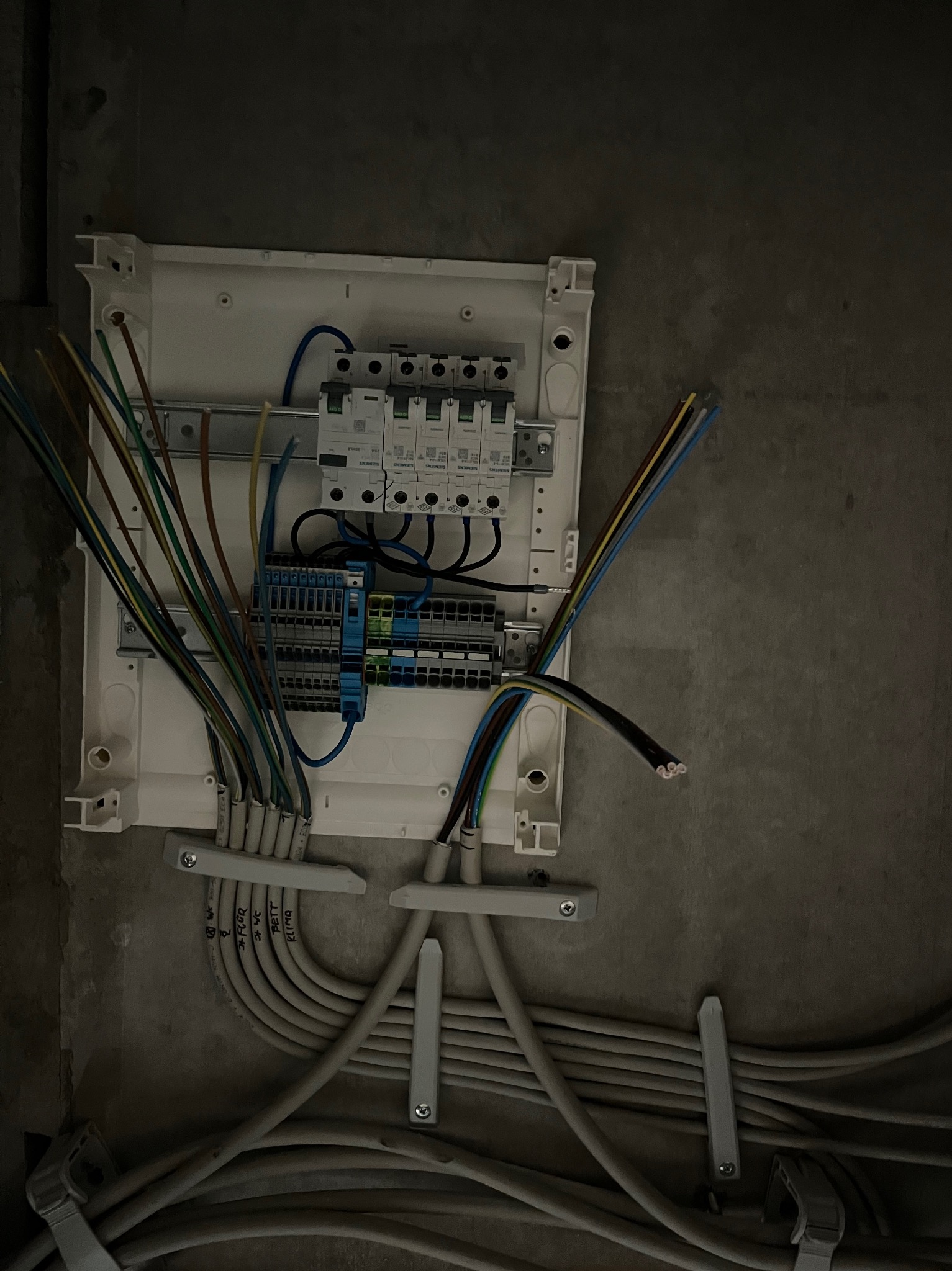 Rozdzielnica elektryczna z podłączonymi przewodami na surowej ścianie. Widoczne zabezpieczenia i listwy montażowe. Przewody opisane. Ujęcie z bliska, ciemne oświetlenie.