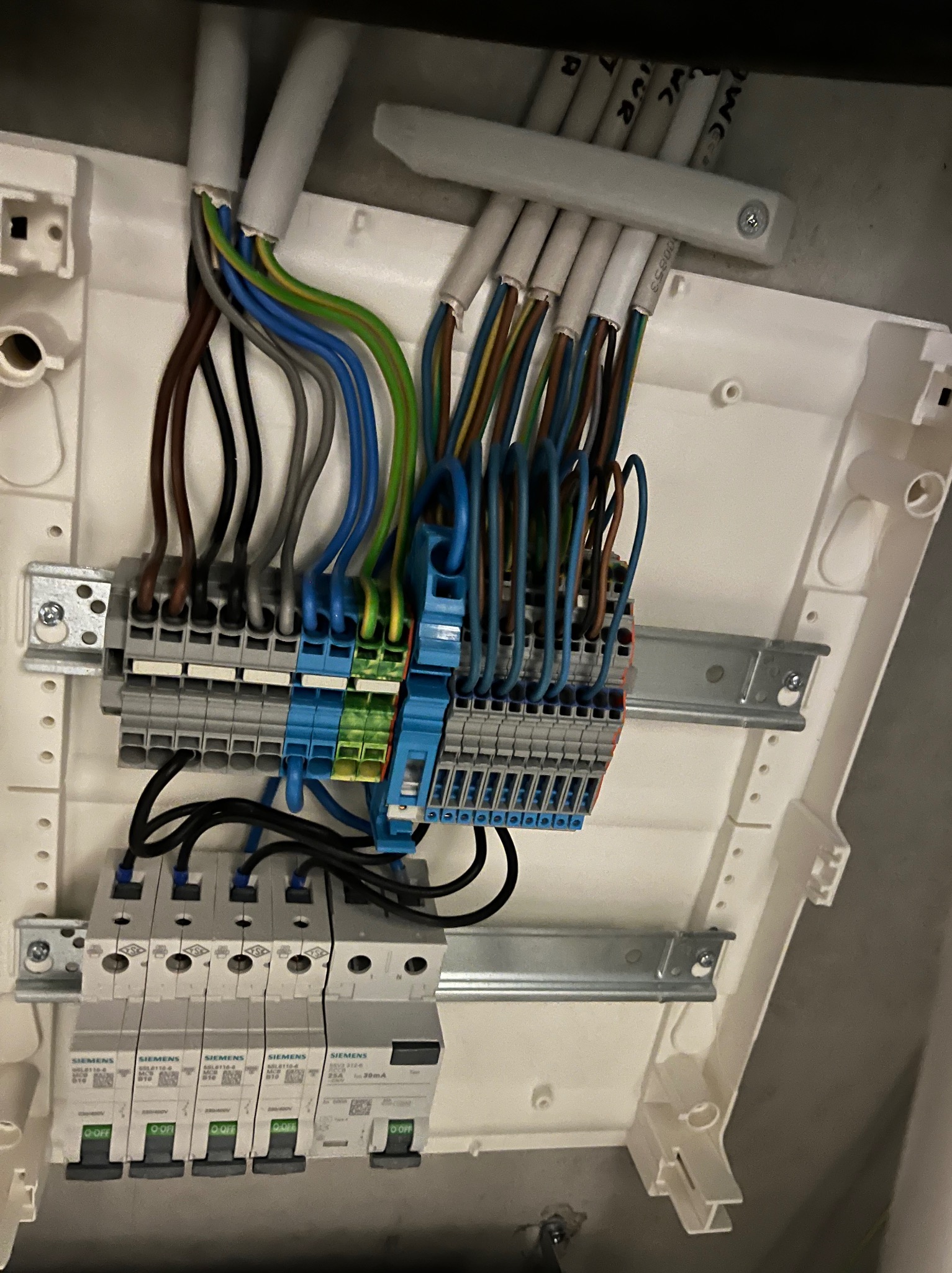 Rozdzielnia elektryczna z podłączonymi przewodami i zabezpieczeniami Siemens. Widoczne kable w różnych kolorach. Instalacja w białej obudowie.