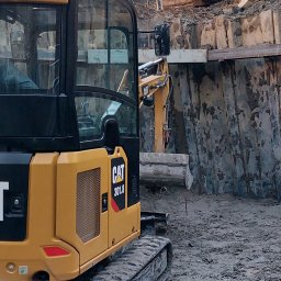 ZHU DARIUSZ PIKÓRA - Żółta koparka CAT 301.8 na placu budowy, widoczne stalowe ścianki wykopu i druga koparka w tle. Detal na gąsienicach i kabinie operatora.