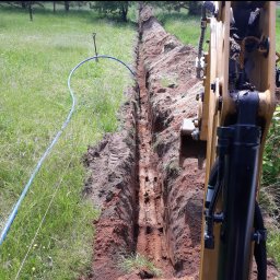 ZHU DARIUSZ PIKÓRA - Wąski wykop w ziemi, z widoczną rurą i fragmentem koparki. W tle zielona trawa i drzewa. Wykop świeżo wykonany, z wyraźnymi śladami pracy maszyny.