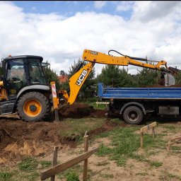 ZHU DARIUSZ PIKÓRA - Żółta koparko-ładowarka JCB nabiera ziemię do niebieskiej ciężarówki na tle zieleni i błękitnego nieba. Widoczne drewniane znaczniki wykopu.