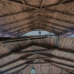 ELASER - Drewniana konstrukcja z widocznymi belkami i lampami, ukazana z perspektywy żabiej. Widoczne elementy krajobrazu w tle. Ciemne drewno, symetryczny układ.
