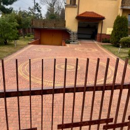 B&L Clean - Kostka brukowa ułożona w okrągły wzór, widoczna zza metalowego ogrodzenia. W tle dom z otwartym garażem i zielenią.