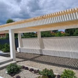 Stachdrew - Nowoczesna pergola z jasnego drewna i białych słupów, rzucająca cienie na żwirowe podłoże. Widok z niskiej perspektywy, zieleń w tle.