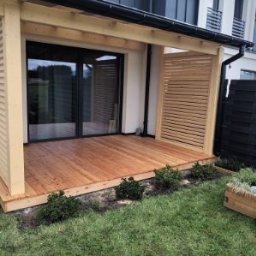 Stachdrew - Drewniany taras z pergolą i lamelami, przylegający do domu z dużym oknem przesuwnym. Widoczne elementy ogrodu i czarne ogrodzenie.