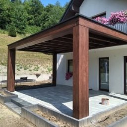 Stachdrew - Ciemnobrązowa pergola z płaskim dachem, przylegająca do białego domu z balkonem. Szara kostka brukowa na tarasie, zielone wzgórze w tle.