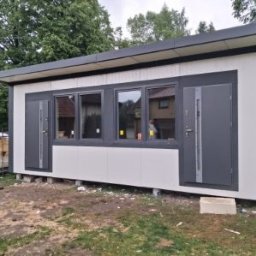 Stachdrew - Nowoczesny, parterowy budynek z płaskim dachem, szare drzwi i okna, białe panele ścienne. Widoczne materiały budowlane obok. Budynek na fundamencie.