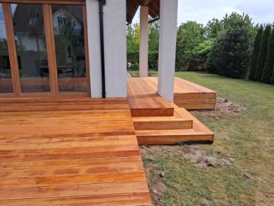 Nowoczesny taras z drewna egzotycznego z niskimi schodami, prowadzący do domu z dużymi oknami. Jasne drewno, zielony trawnik i minimalistyczna architektura.