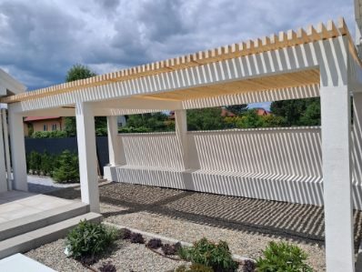 Nowoczesna pergola z jasnego drewna i białych słupów, rzucająca cienie na żwirowe podłoże. Widok na ogród i dom w tle, słoneczny dzień.