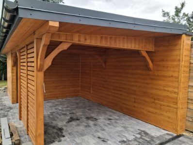 Drewniana konstrukcja z zadaszeniem i ażurowymi ścianami bocznymi, wykończona w ciepłym odcieniu brązu, na tle szarej kostki brukowej. Widoczne elementy dekoracyjne z drewna.