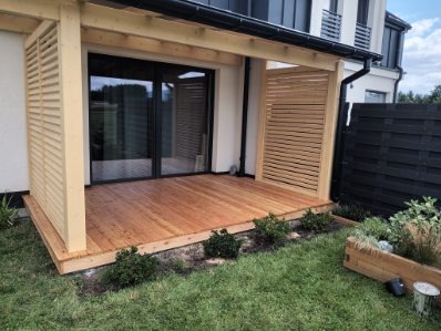 Drewniany taras z pergolą i lamelami, przylegający do domu z dużym oknem przesuwnym. Widoczne elementy ogrodu i czarne ogrodzenie.